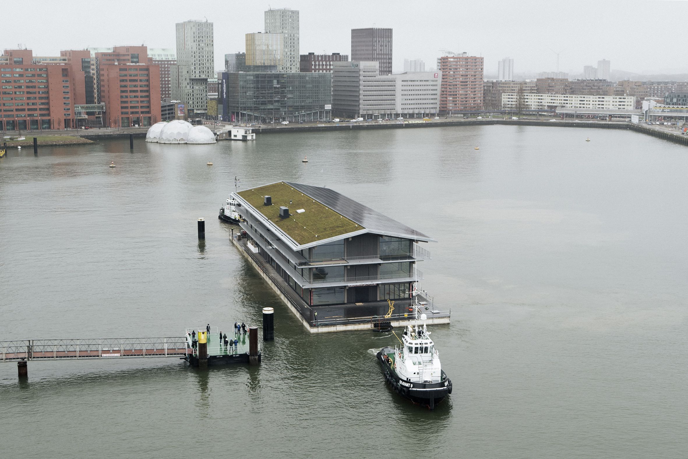 Im Rotterdamer Hafenbecken (Rijnhaven) wird das „Floating Office“ per Schlepper an seine Position gebracht. Dabei ist das Satteldach halbseitig begrünt und halbseitig mit Photovoltaikmodulen belegt.